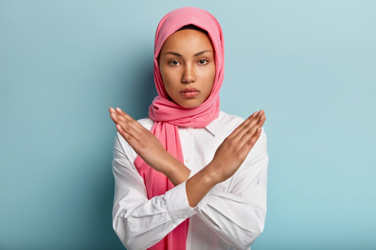 Beautiful serious Arabian woman wears pink hijab, makes cross with hands over chest, shows stop or prohibition gesture, disagrees with uninteresting offer, isolated over blue background. No sign