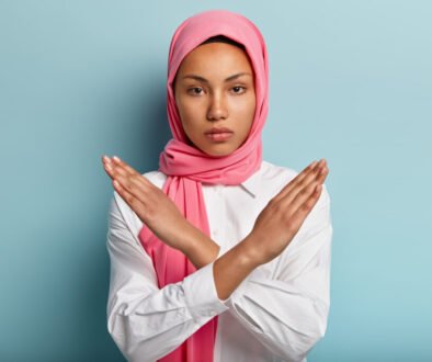 Beautiful serious Arabian woman wears pink hijab, makes cross with hands over chest, shows stop or prohibition gesture, disagrees with uninteresting offer, isolated over blue background. No sign