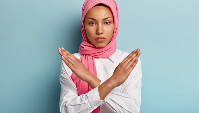 Beautiful serious Arabian woman wears pink hijab, makes cross with hands over chest, shows stop or prohibition gesture, disagrees with uninteresting offer, isolated over blue background. No sign